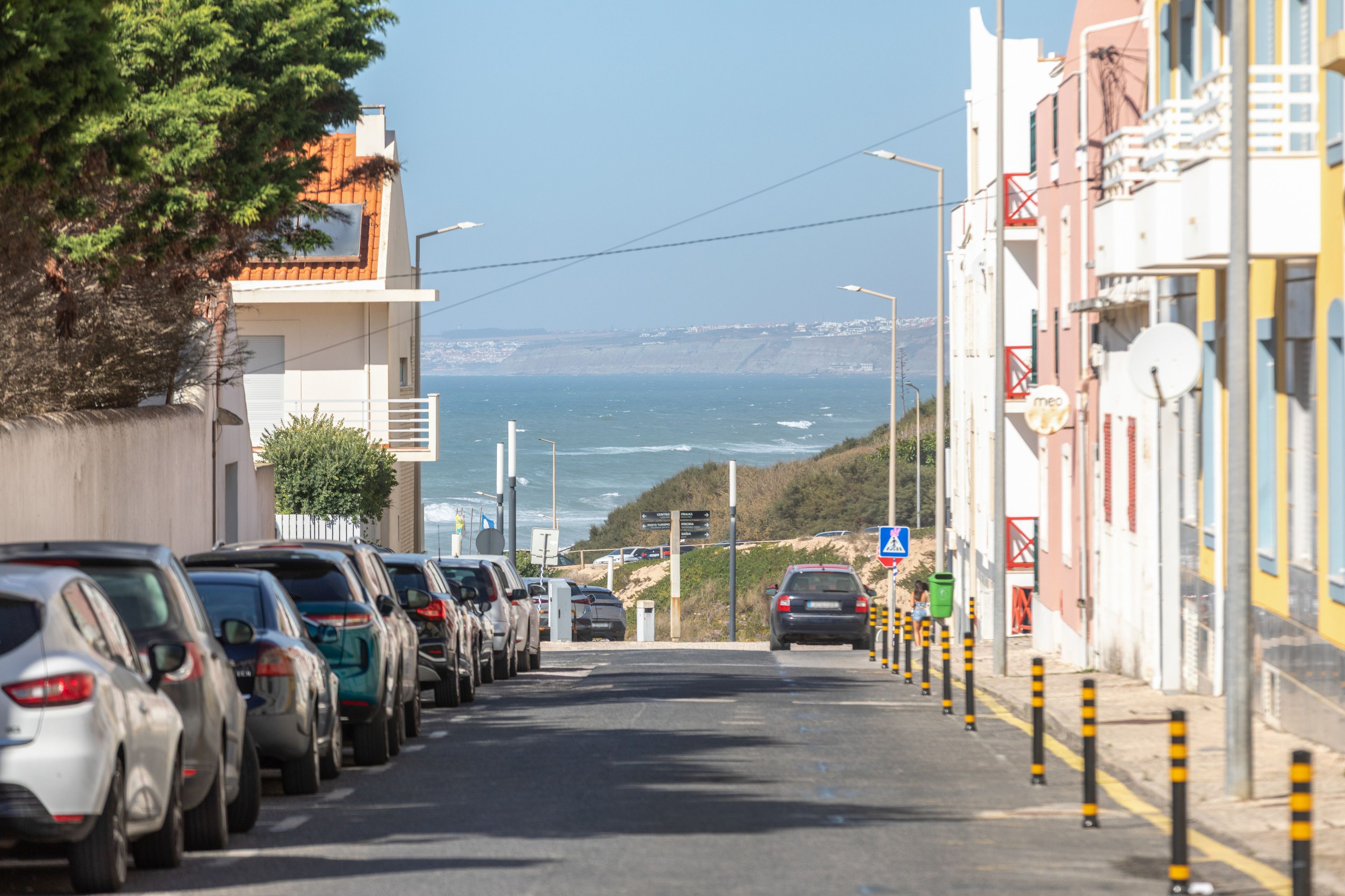 A coastal street lined with parked cars and buildings, leading towards the sea.