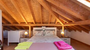 Quarto com teto inclinado, vigas de madeira, cama com roupa de cama floral, duas luminárias de cabeceira e um radiador.