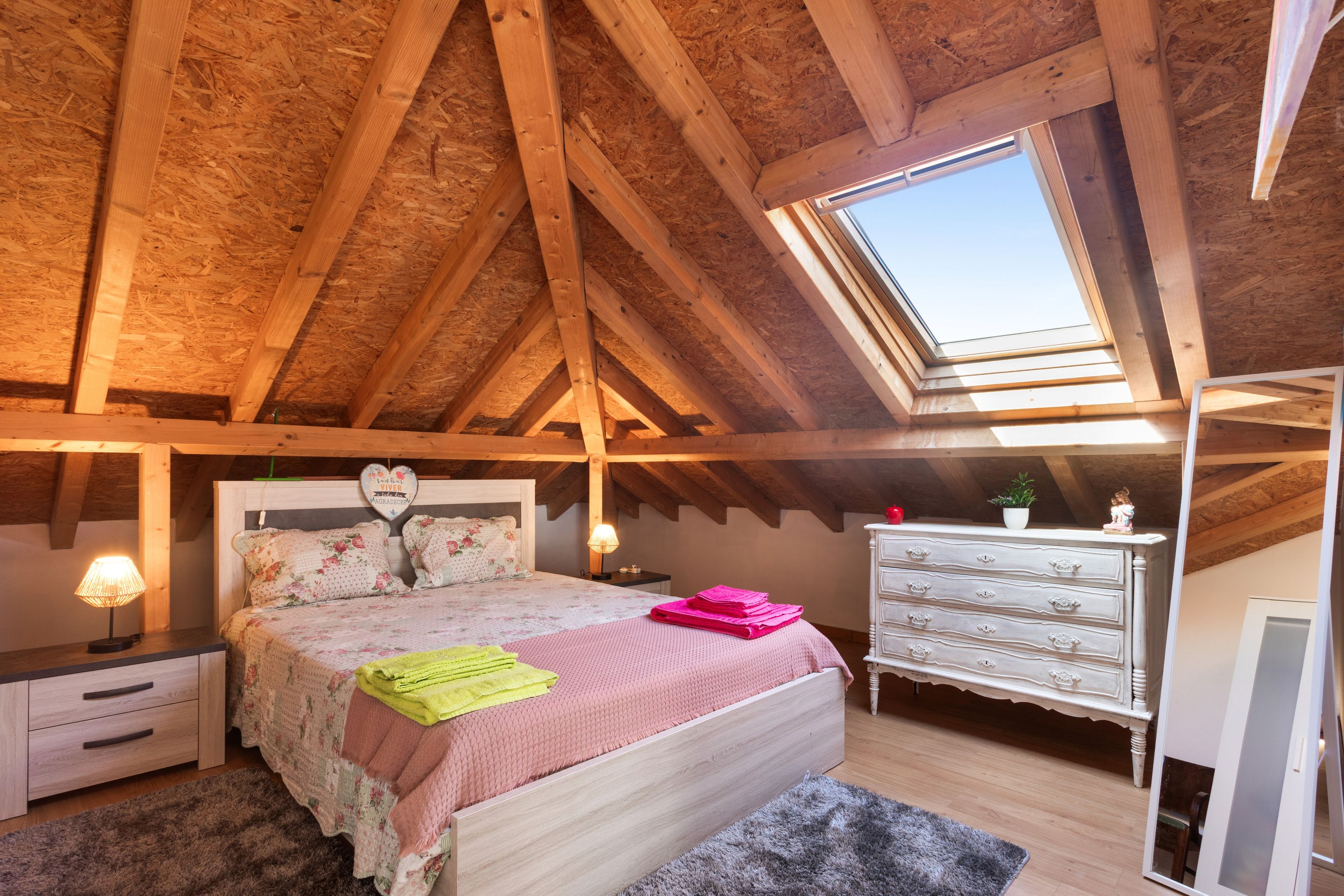 Una camera da letto con soffitto spiovente, travi in legno, un lucernario, un letto con lenzuola a motivi floreali, un comò e un comodino con una lampada.