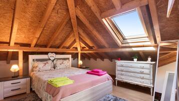 Una camera da letto con soffitto spiovente, travi in legno, un lucernario, un letto con lenzuola a motivi floreali, un comò e un comodino con una lampada.