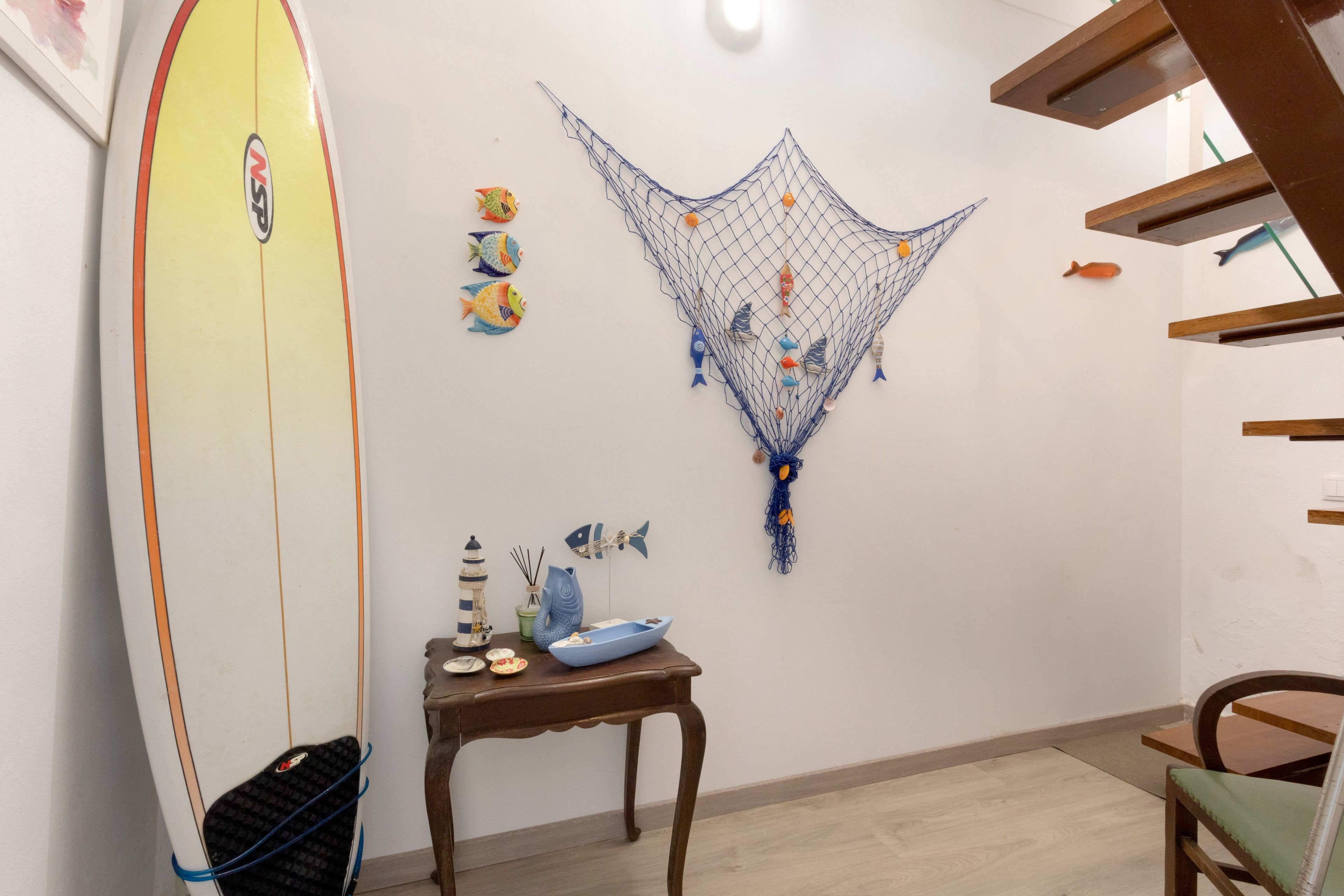 Ein Zimmer mit einem an der Wand liegenden Surfbrett, einem kleinen Tisch mit Dekoration und einer Wandgestaltung mit Fischen und Fischernetzen.