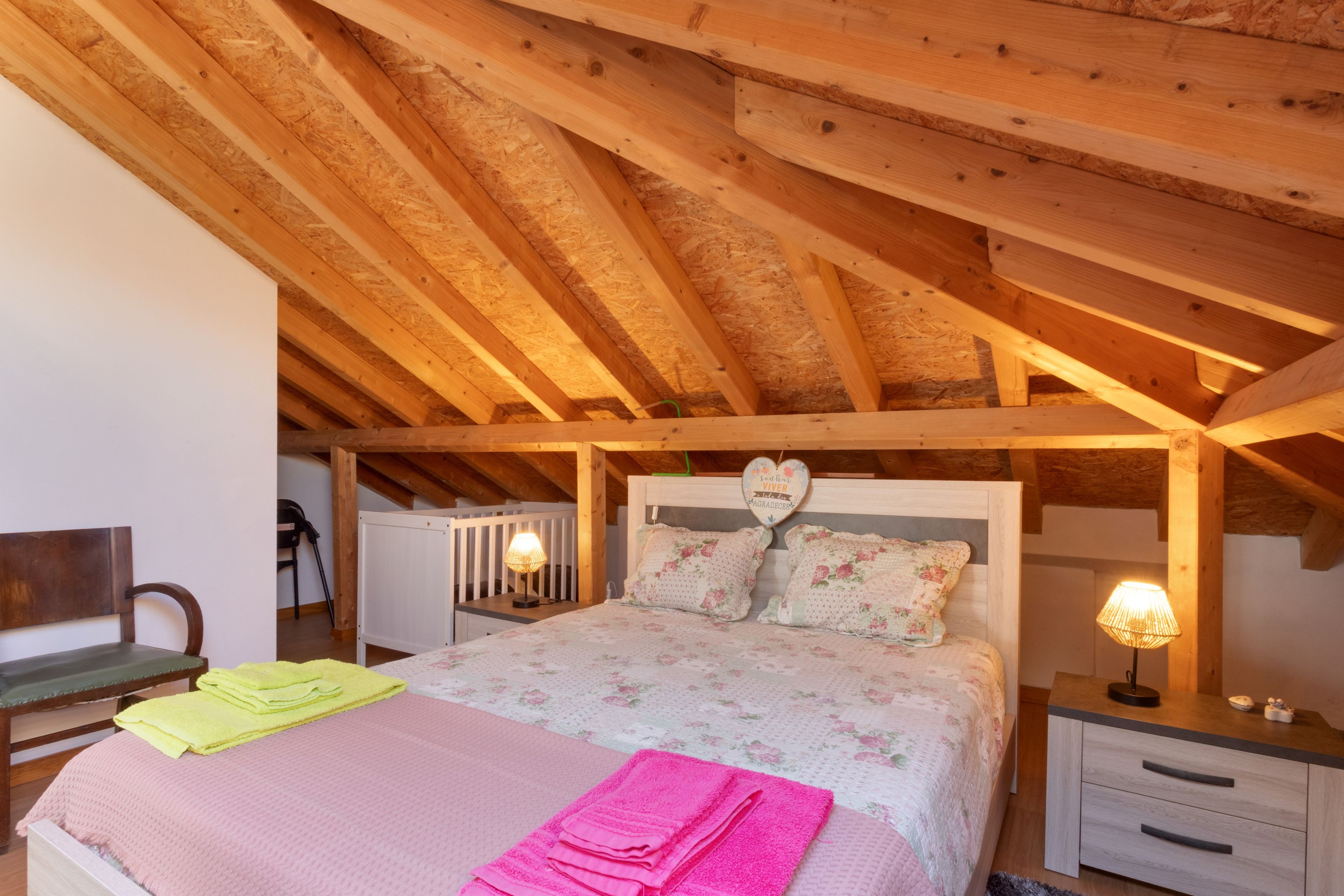Quarto com teto de madeira, cama com roupa de cama florida, duas luminárias de apoio e decoração em formato de coração na cabeceira.