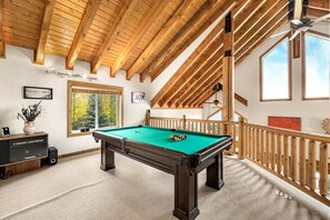 Une table de billard dans une pièce avec des poutres en bois, une cheminée et une vue sur les arbres par la fenêtre.