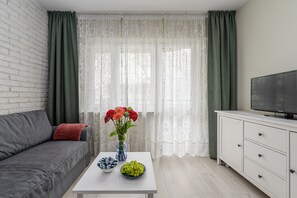 A living room with a grey sofa, a white coffee table with a vase of flowers, a bowl of grapes, and a television on a white cabinet.