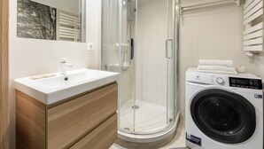 Un baño moderno con ducha envuelta en vidrio, lavamanos con tocador de madera y lavadora de carga frontal.