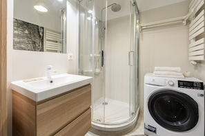 A modern bathroom with a glass-enclosed shower, a sink with a wooden vanity, and a front-loading washing machine. - Wileńska P&O Apartments (Warszawa)