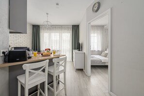 A modern kitchen with a dining area, a small white cabinet, and a bed in the adjacent room.