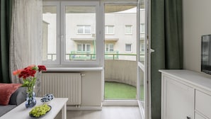 Una habitación con vistas a un edificio a través de una ventana, un radiador, una mesa con flores y un frutero, y un televisor sobre un mueble.
