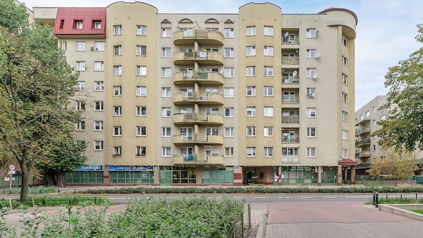 Un edificio residencial de varios pisos con balcones, una sección en esquina con tejado rojo y un área pavimentada con zonas verdes en la parte frontal.
