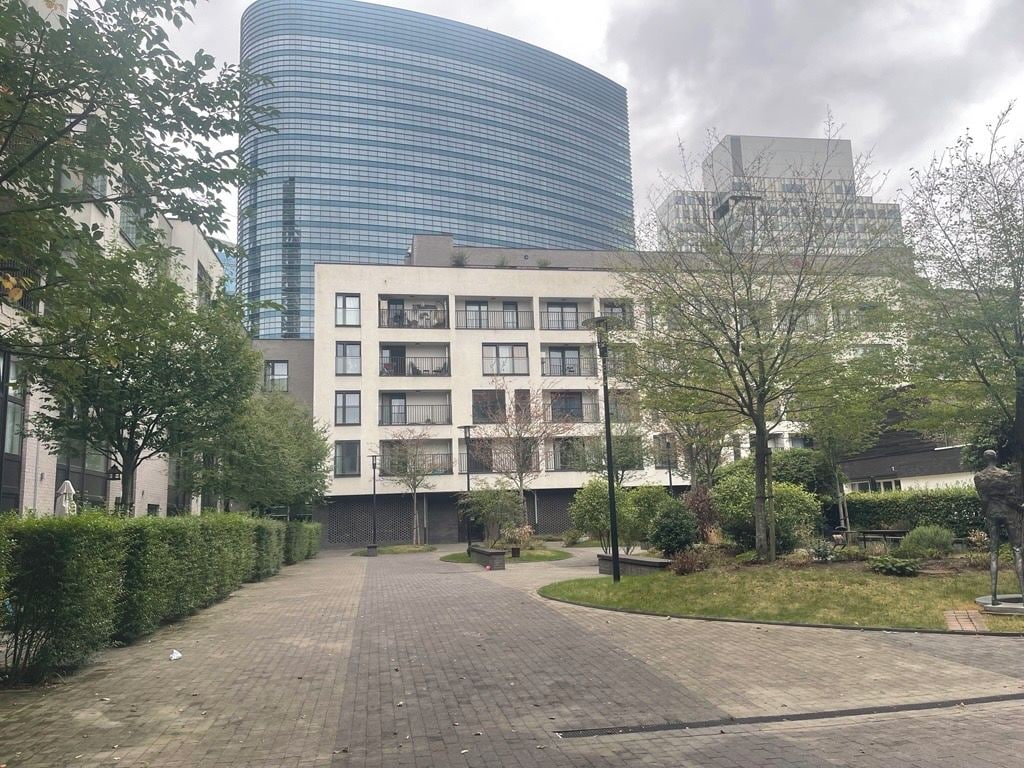 Een gebouw met een gebogen glazen gevel, een plat dak met meerdere ramen, en een geplaveid binnenhof met bomen en een standbeeld.
