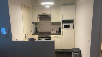Een kleine keuken met witte kastjes, een magnetron, een kookplaat en een prullenbak.