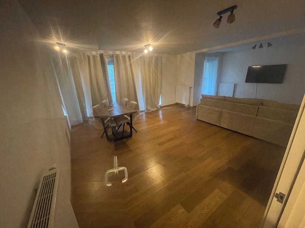 Een ruime woonkamer met een houten vloer, een bank, een eettafel met stoelen en een aan de muur bevestigde televisie.