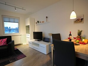 Un salon moderne avec un téléviseur, un meuble de rangement blanc et un coin repas comprenant une table et des chaises en bois.