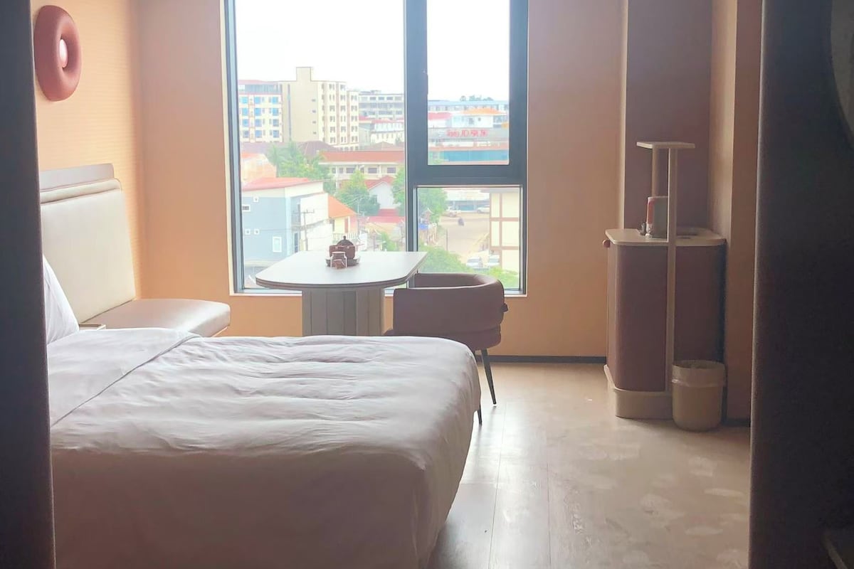 Habitación de hotel con cama, una mesita, una silla y vistas a edificios a través del ventanal.