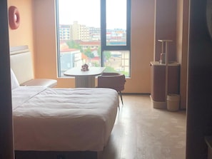 Quarto de hotel com cama, uma mesinha, uma cadeira e vista para edifícios pela janela.