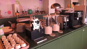 Una zona de cocina con diversos electrodomésticos, incluyendo un espumador de leche, una cafetera y una licuadora. También hay pilas de platos y una bandeja con moldes para muffins.