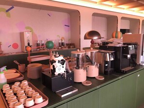 Cozinha equipada com diversos eletrodomésticos, incluindo um espumador de leite, máquina de café e liquidificador. Há também pilhas de pratos e uma bandeja com forminhas para muffins.