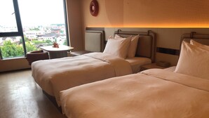 Habitación de hotel con dos camas, una mesita pequeña y vista a la ciudad a través de amplios ventanales.
