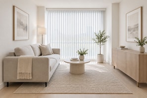 Una sala de estar moderna con un sofá color beige, una mesa de centro redonda, una planta en maceta, un mueble de madera y un ventanal con persianas.