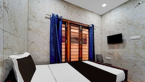 A bedroom with a bed, a window with curtains, a television mounted on the wall, and a power outlet. - Townhouse Gandhipuram Central Bus Stand (Coimbatore)