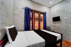 Un dormitorio con cama, ventana con cortinas, televisor montado en la pared y toma de corriente.