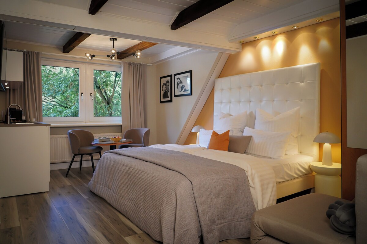 A bedroom with a large bed, a desk, and a chair. There is a window with curtains, a ceiling fan, and a wall-mounted lamp.