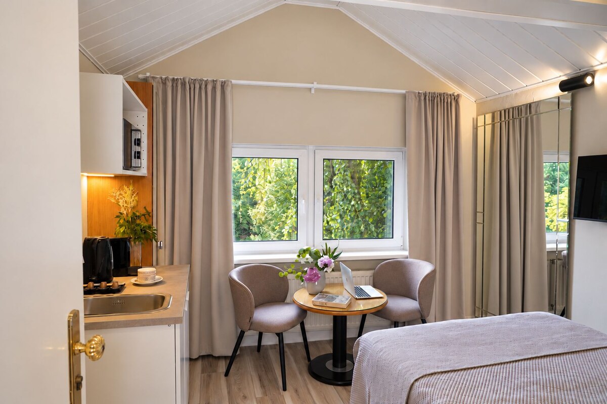 A room with a bed, a small table with a laptop, two chairs, a window with curtains, and a kitchenette area.