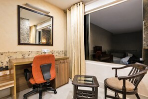 A hotel room with a desk, two chairs, a large window with curtains, and a mirror on the wall.