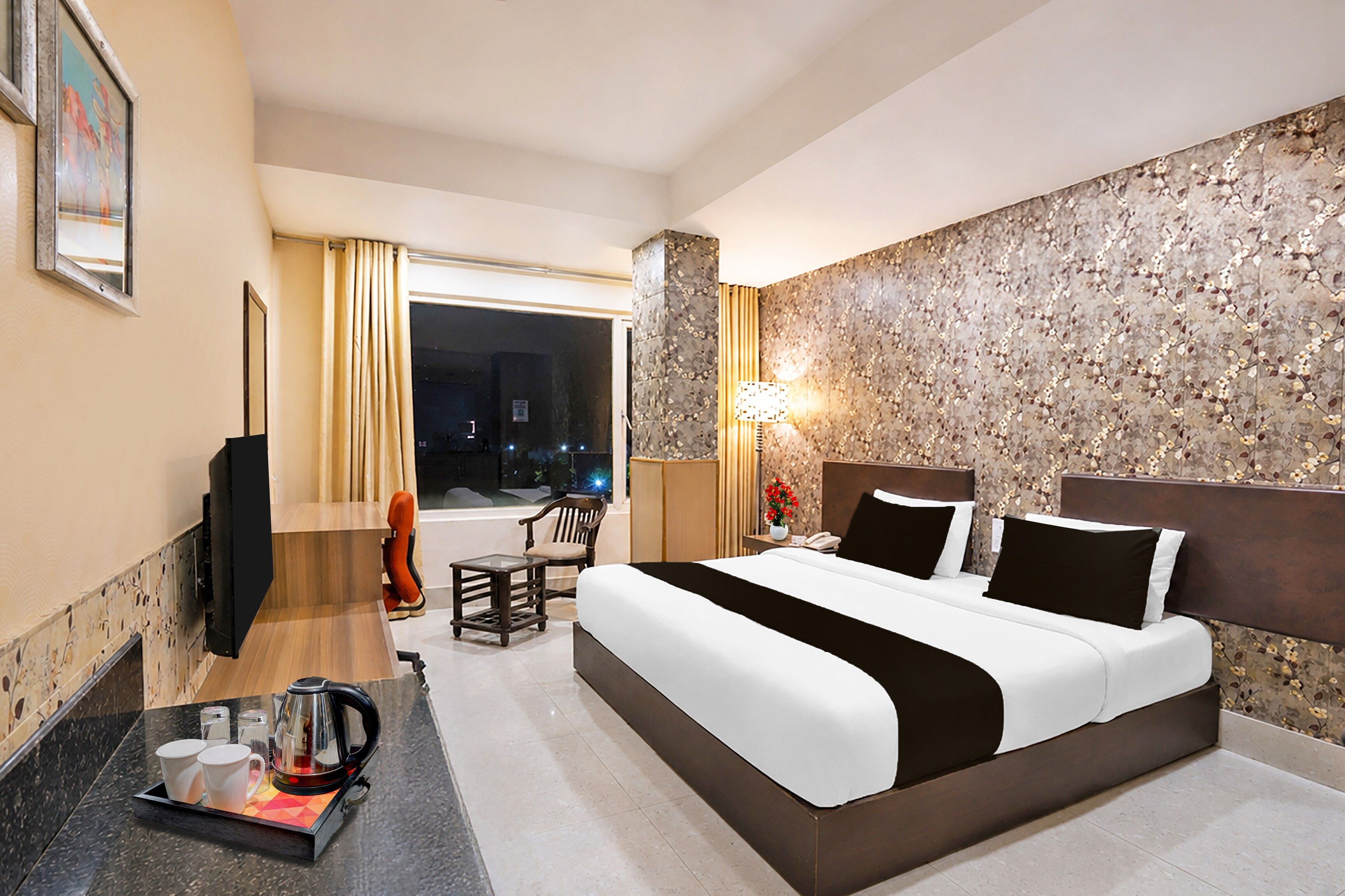 A hotel room with a large bed, a desk with a kettle, a chair, and a view of the city at night.