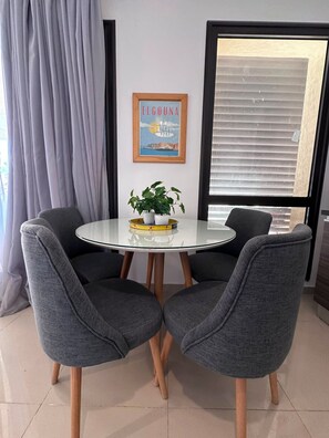 A dining area with a round glass table, two grey upholstered chairs, a potted plant, and a framed picture on the wall.