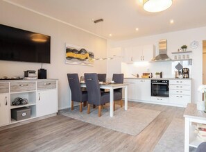 A modern kitchen with white cabinetry, a dining area with a table and chairs, and a flat-screen TV mounted on the wall. - Sea View Villa, Apartment 9 "beach Love" (Grossenbrode)