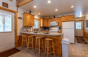 A kitchen with wooden cabinets, a central island with stools, a microwave, stove, and a refrigerator. - Cozy Cabin Retreat with Hot Tub & Fire Pit for Summer in Island Park, Idaho (Island Park)