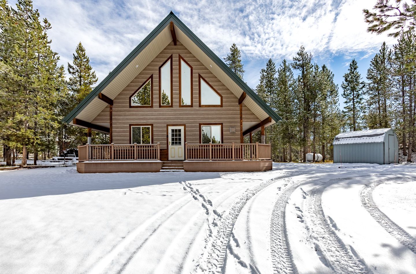 A cabin with a prominent triangular roof, a porch with railings, and a snow-covered ground with tire tracks.