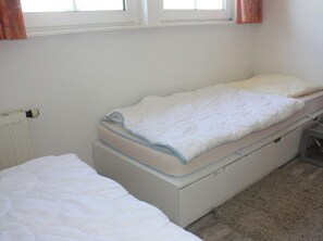 A single bed with a white mattress and blue headboard in a room with a radiator and a window with curtains. - Holiday Apartment With 1 Bedroom (Cuxhaven)