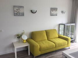 Una sala de estar con un sofá amarillo, una mesita de noche blanca con lámpara y un mueble con puertas de vidrio.