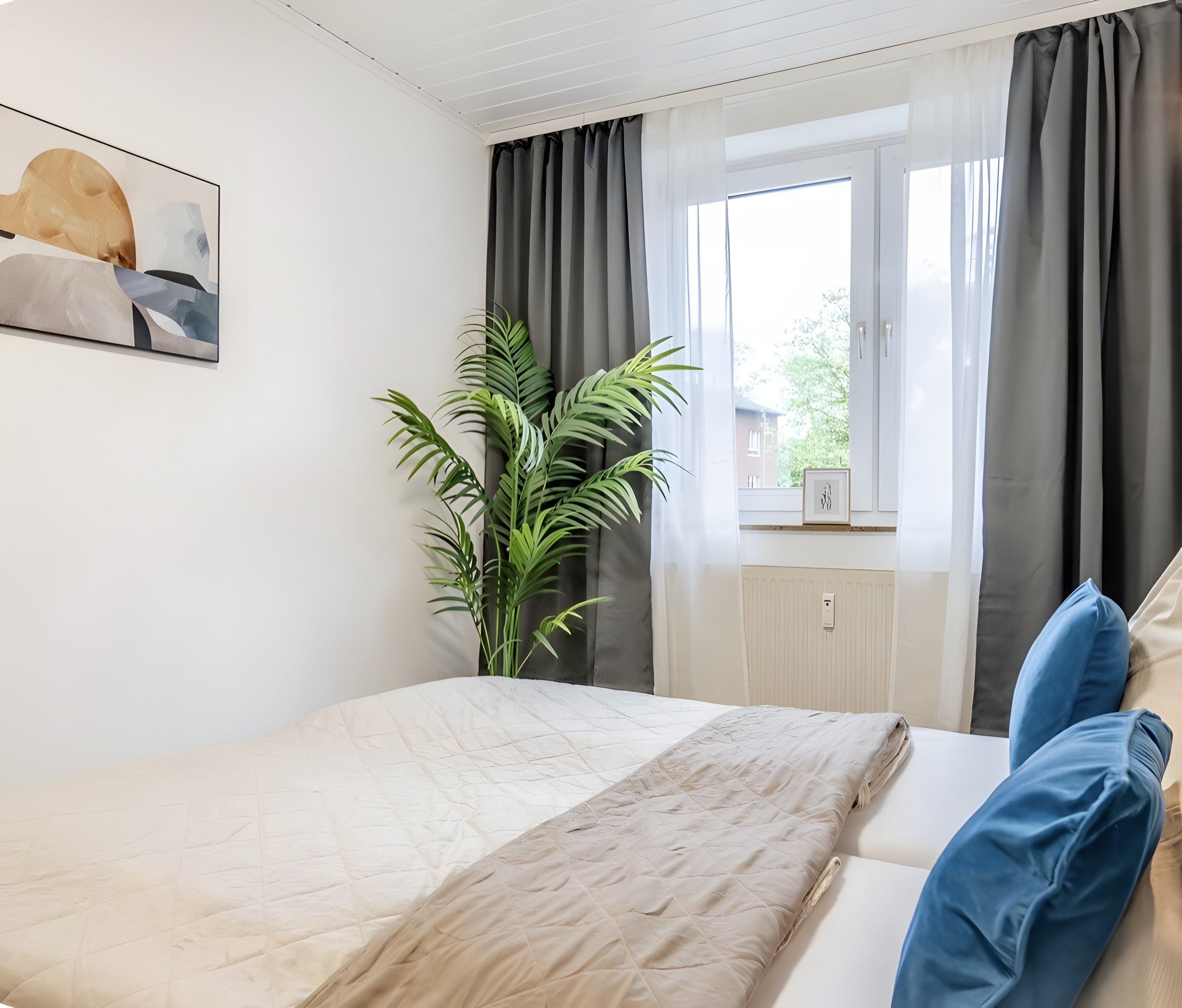 Ein Schlafzimmer mit einem Bett, einer Topfpflanze, einem Fenster mit Vorhängen und einem Wandbild.