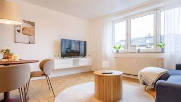Una sala de estar moderna con un televisor de pantalla plana, una mesa de centro redonda de madera, un sofá, una mesa de comedor con sillas y una ventana con cortinas.