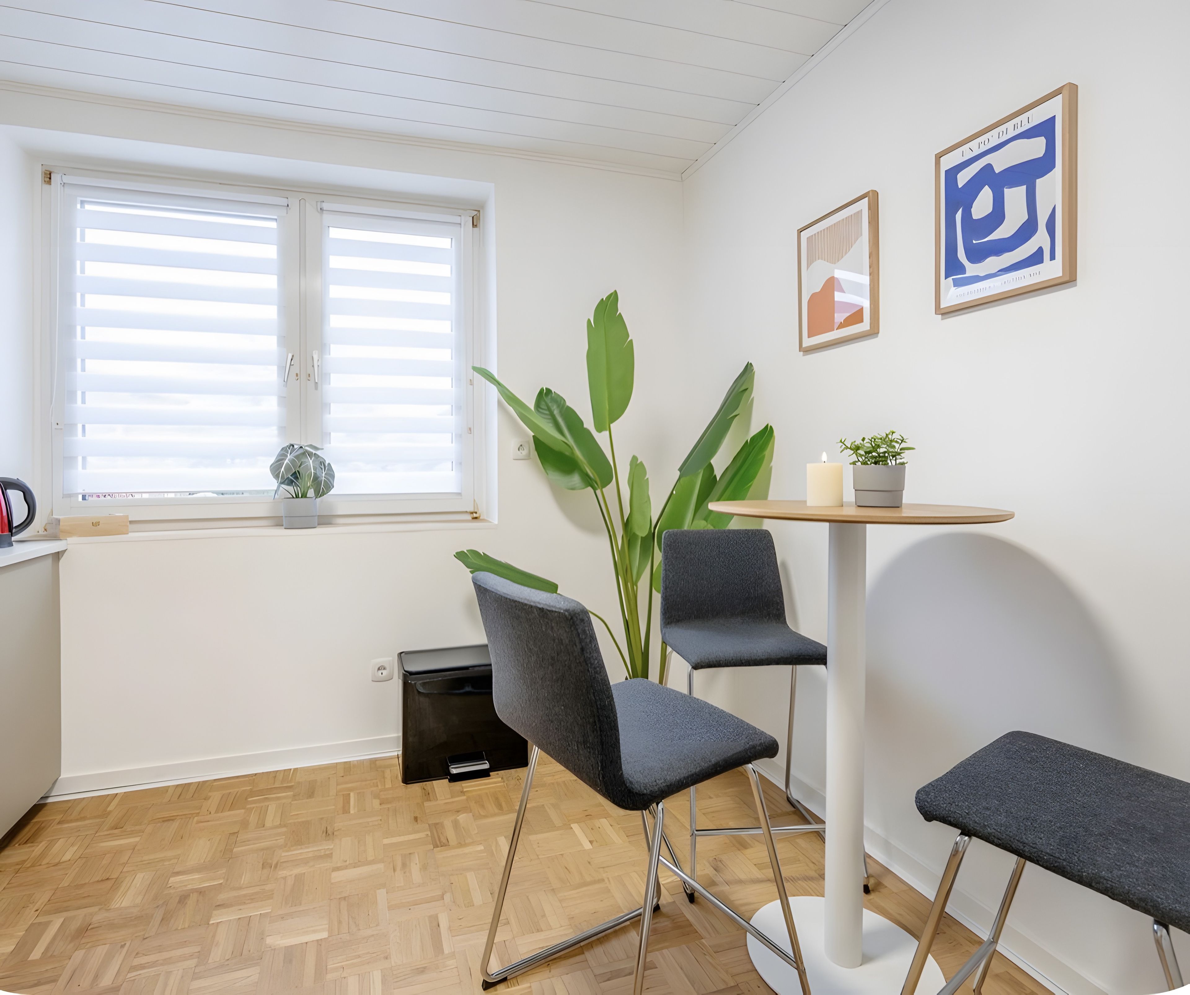 Ein modernes Zimmer mit Holzboden im Fischgrätmuster, einem runden Tisch mit zwei Stühlen, einer Topfpflanze und zwei gerahmten Kunstwerken an der Wand.