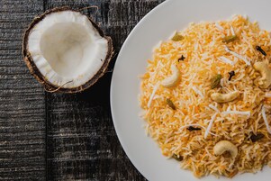Un plato de arroz amarillo con pasas y nueces de anacardo, junto a un coco partido sobre una superficie de madera oscura.