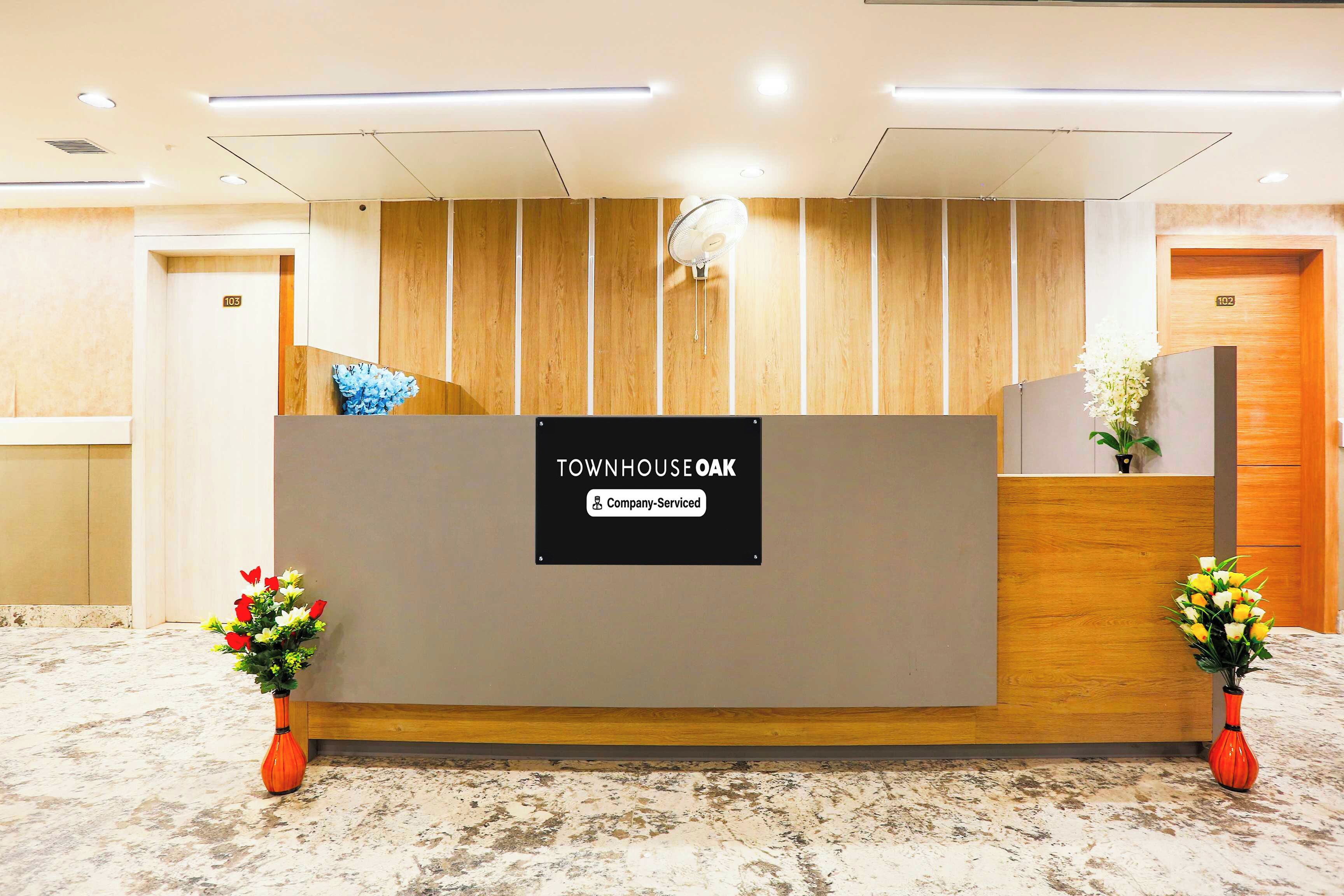 A reception desk with a sign indicating it's company-serviced, flanked by two flower arrangements, and a wooden panel wall behind.