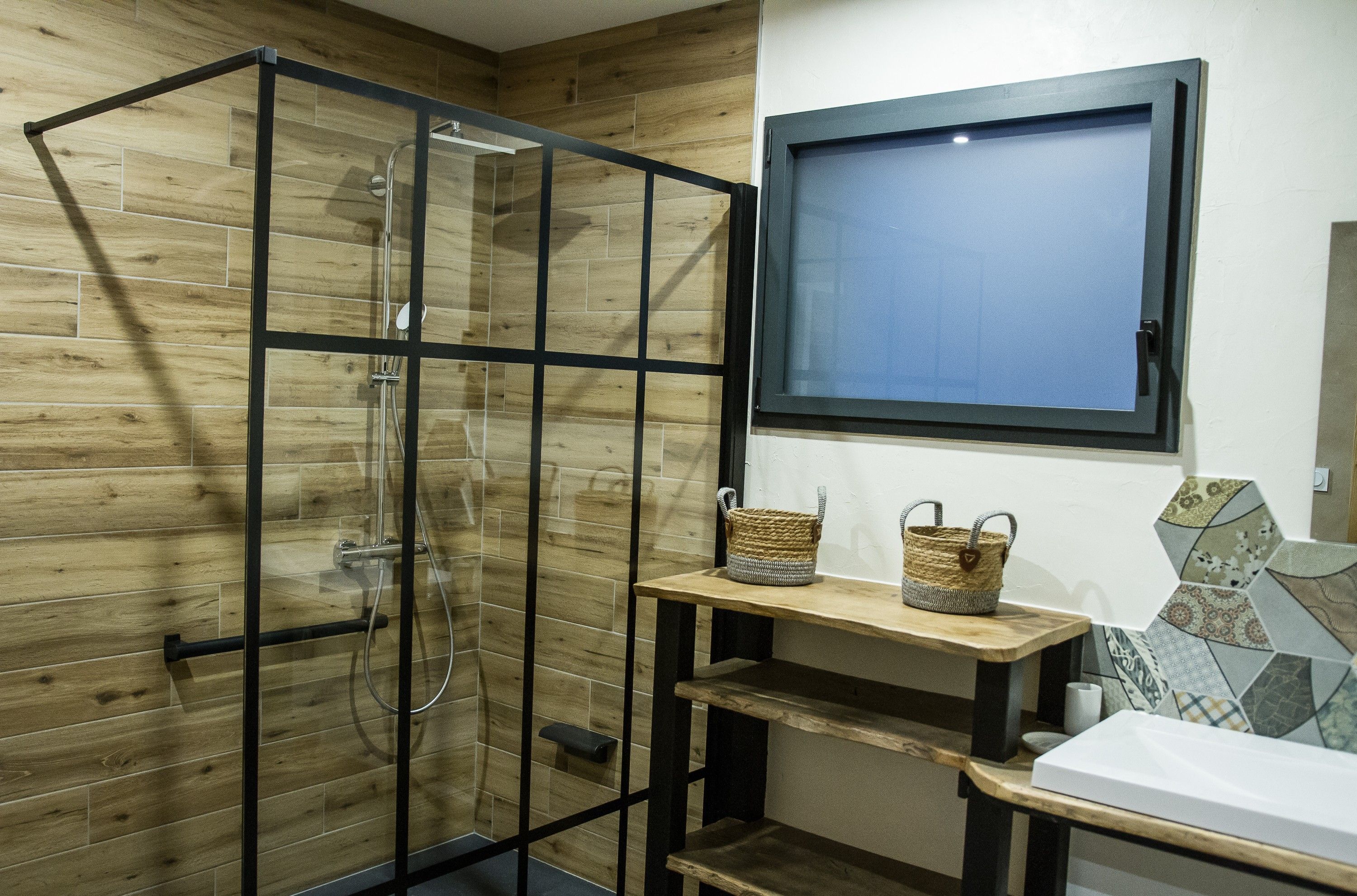 Une douche avec des portes en verre, un mur en bois, une étagère avec des paniers et une fenêtre donnant sur un paysage bleu.