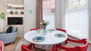 Un comedor con una mesa redonda, sillas rojas, un jarrón con flores y una estantería con libros y objetos decorativos.