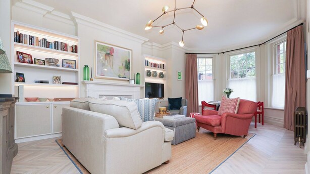 Una sala de estar con sofá, sillón, reposapiés, chimenea, estanterías y un gran ventanal con cortinas.