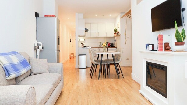 Una sala moderna con sofá, comedor con mesa y sillas, cocina con armarios, televisor montado en la pared y chimenea.