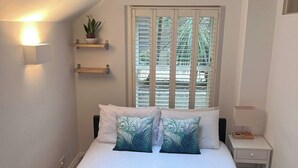 Un dormitorio con cama, almohadas, una mesita de noche, estante en la pared con una planta y un tragaluz.