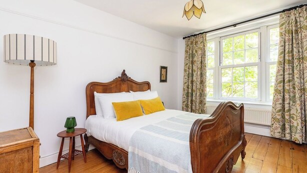 Un dormitorio con cama de madera, lámpara de noche, una mesita y una ventana con cortinas.