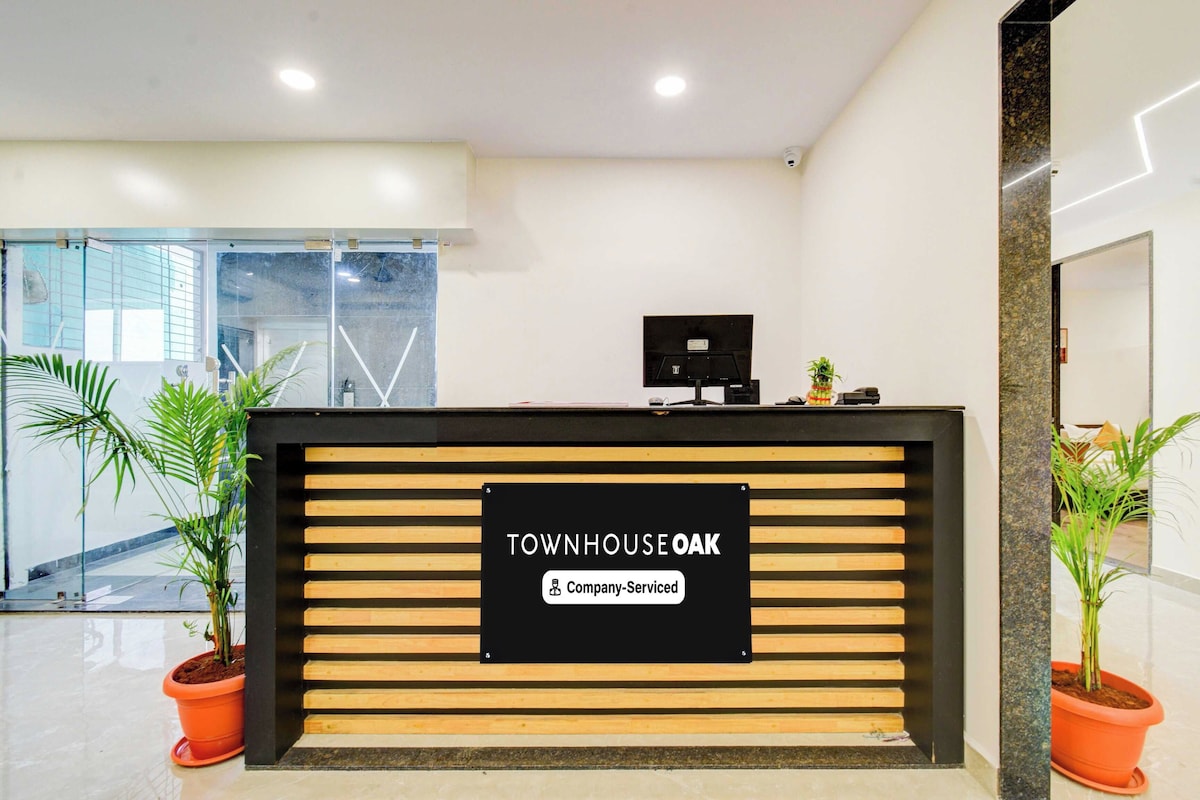 Un mostrador de recepción con un cartel que dice 'TOWNHOUSE OAK' y 'Servicio de empresa'. Hay plantas en macetas a ambos lados del mostrador.