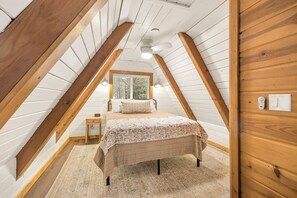A bedroom with a sloped ceiling, a bed with a floral bedspread, a nightstand, and a window with a view of trees. - Inviting Riverside Vacation Home with Private Dock Access near Island Park, Idaho (Island Park)