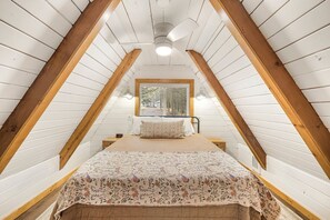 Ein Schlafzimmer mit schrägen Wänden, einem Bett mit Blumenmuster auf der Bettwäsche, einem Nachttisch und einem Fenster mit Blick auf Bäume.