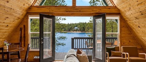 Una cabaña acogedora con interior de madera, un sofá, sillas y vista a un lago a través de puertas abiertas.